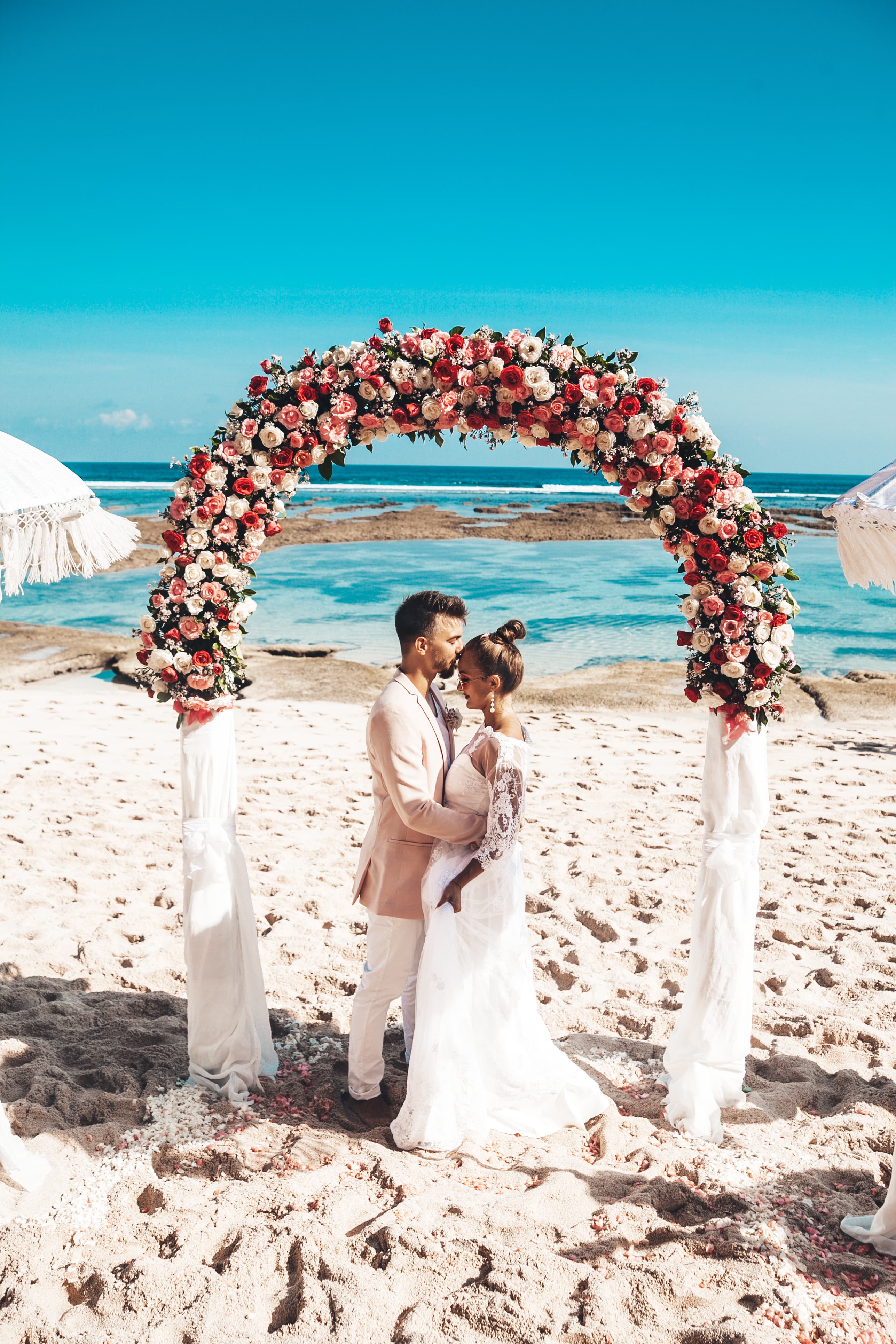 Pareja de recién casados abrazándose bajo un arco de flores en una boda en la playa, con el mar de fondo.